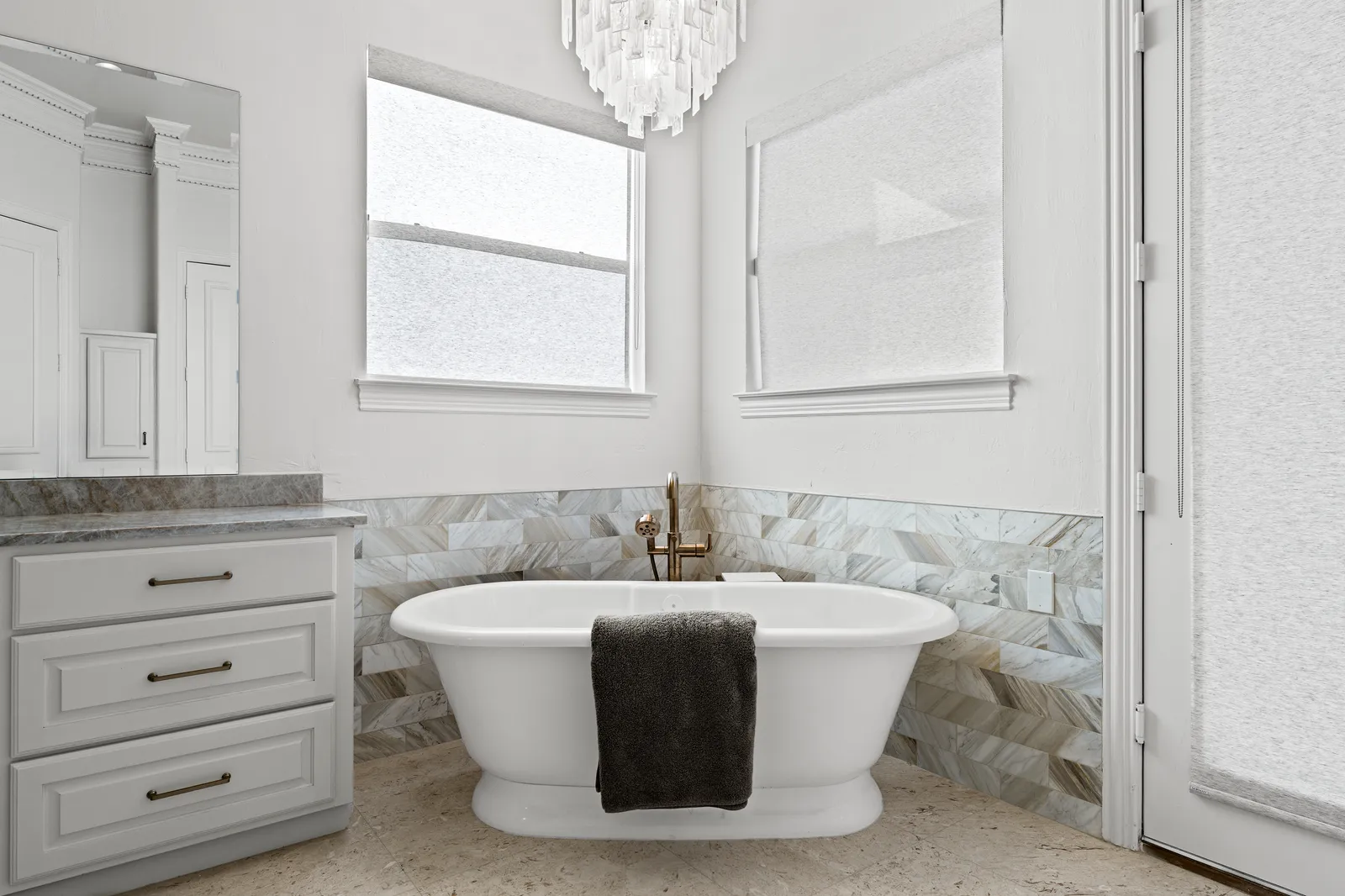 Primary bathroom at Prestwick Ln in Dallas after the renovation: freestanding tub, chevron marble-look tile wainscot, brass tub filler, and a crystal chandelier