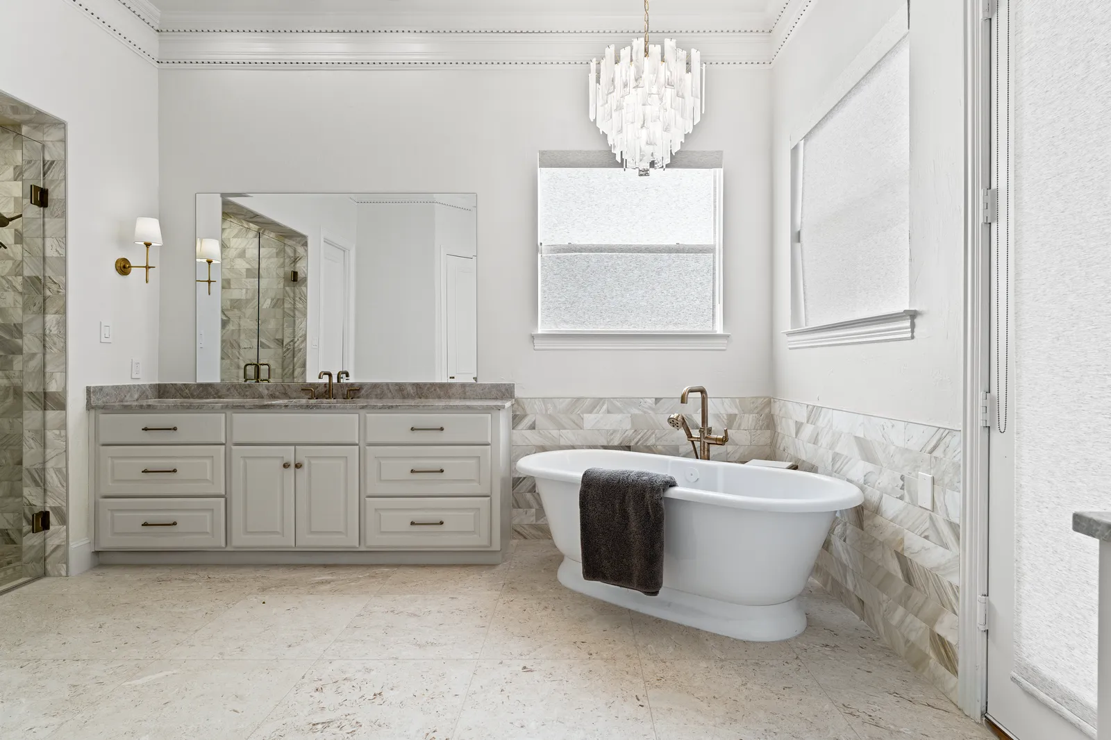 Primary bathroom at Prestwick Ln in Dallas after the renovation: painted vanity with stone counters, freestanding tub, chevron tile wainscot, and a chandelier