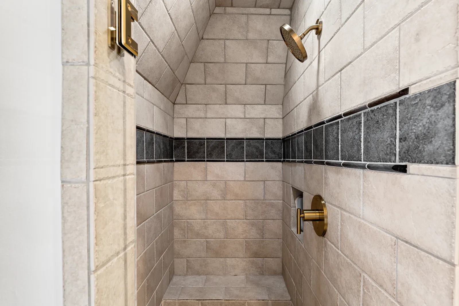 Guest bath shower at Prestwick Ln in Dallas after the renovation: beige tile with a dark accent band and brass fixtures